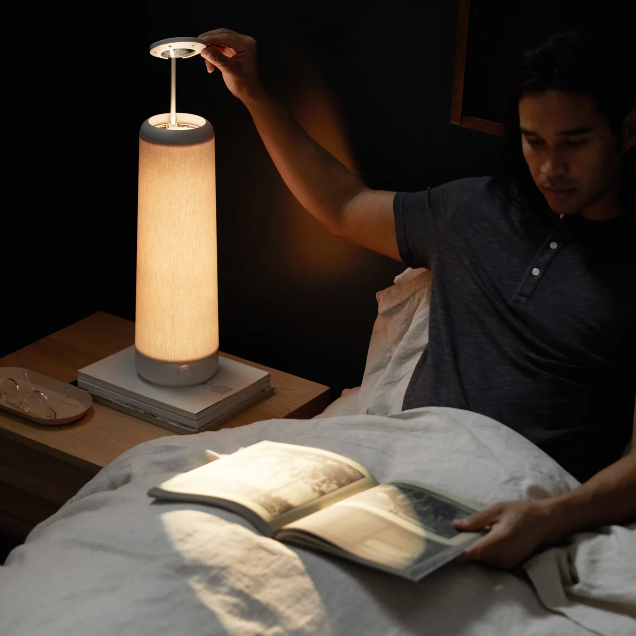 A man in bed adjusts his Nightside NS01 lamp while reading a book. The room is dimly lit, and a pair of glasses rests on a nearby nightstand. The person seems relaxed and focused on the book.