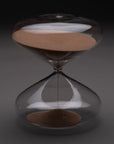 The Intelligent Change Mindful Focus 30 Minute Hourglass, featuring brown sand mostly in the upper half, stands upright on a gray surface with a dark background, representing mindfulness and effective time management.