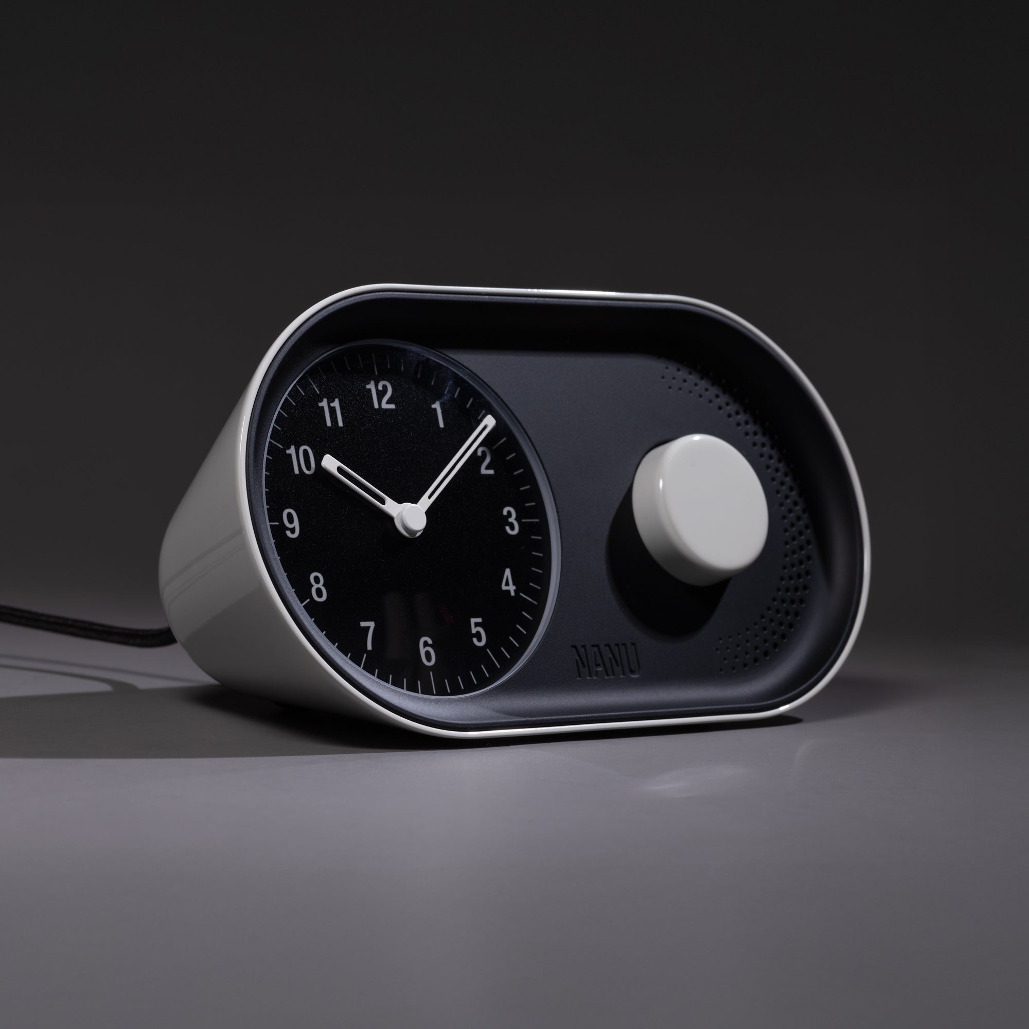 The Nanu Arc Alarm Clock features a modern black and white analog design with a circular dial and large knob, sitting on a smooth surface and casting a shadow in minimal lighting. Battery backup ensures uninterrupted timekeeping.