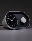 The Nanu Arc Alarm Clock features a modern black and white analog design with a circular dial and large knob, sitting on a smooth surface and casting a shadow in minimal lighting. Battery backup ensures uninterrupted timekeeping.