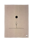 The Midnight Sun Throw Blanket by Blacksaw features a minimalist beige fabric made from eco-friendly fibers, a black vertical line with two branches below a solid black circle like a sun or moon, and includes a small white label in the bottom right corner.