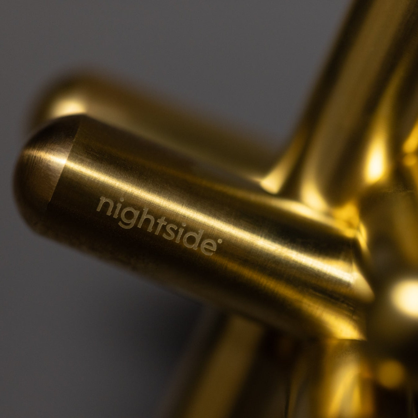 Close-up of intersecting gold metal rods with nightside engraved on one, set against a softly blurred gray background, showcasing sleek modern design.