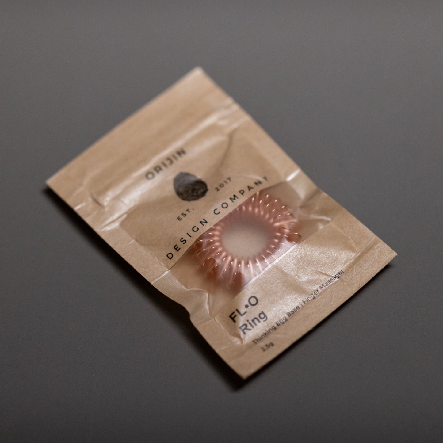 A small brown paper package labeled DESIGN COMPANY holds a pink, coiled ring-shaped accessory, possibly a hair tie, set against a plain gray background.