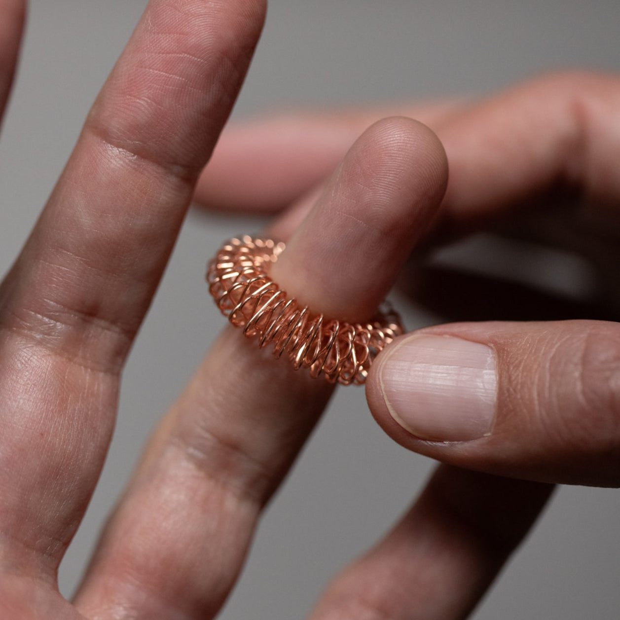 A close-up of a hand slipping a metallic acupressure massage ring onto a finger, with another hand steadying the ring for gentle pressure against a neutral background.