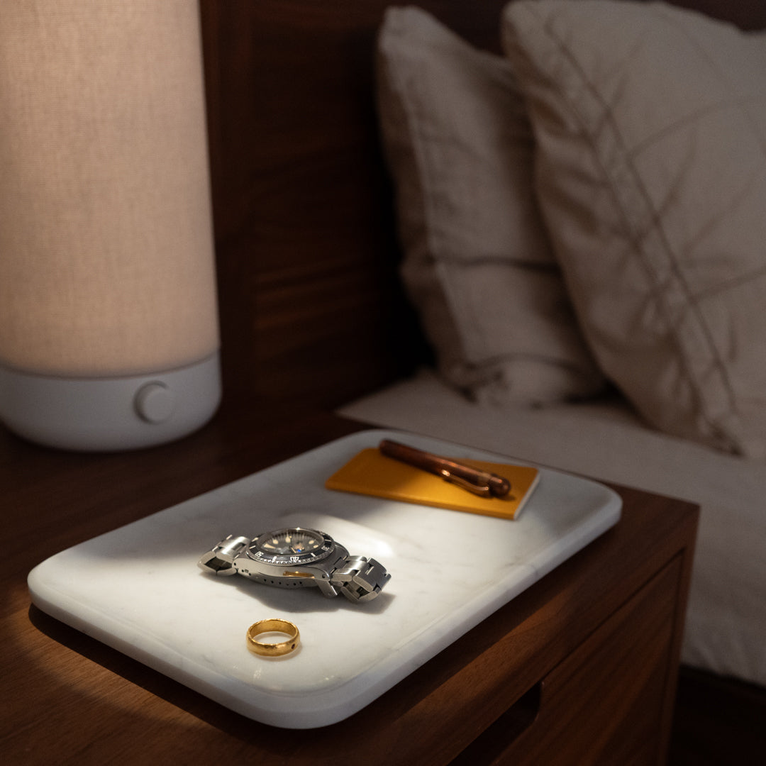 A bedside scene with a sleek table lamp, a marble tray holding a wristwatch, a gold ring, and a bronze pen on a yellow notepad, next to a bed with white pillows. The lighting creates a cozy and intimate atmosphere.