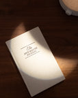 An undated Five Minute Journal by Intelligent Change rests on a wooden surface, partially lit by a circular light. It has a plain, light-colored cover with the title in bold black letters and includes 264 pages for six months of mindful reflection.
