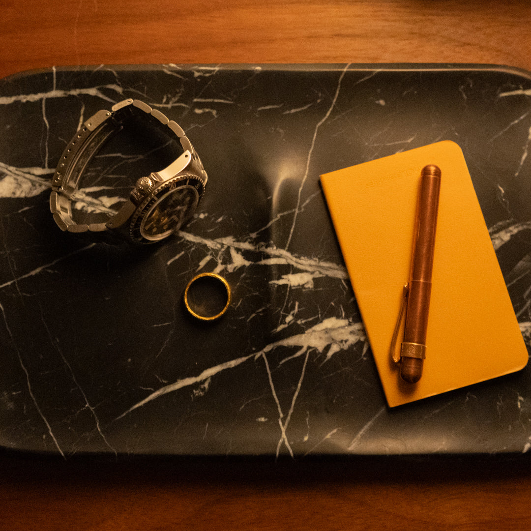A rectangular black marble tray holds a silver watch, a gold ring, and a small yellow notebook with a brown pen on top, all set on a wooden surface.