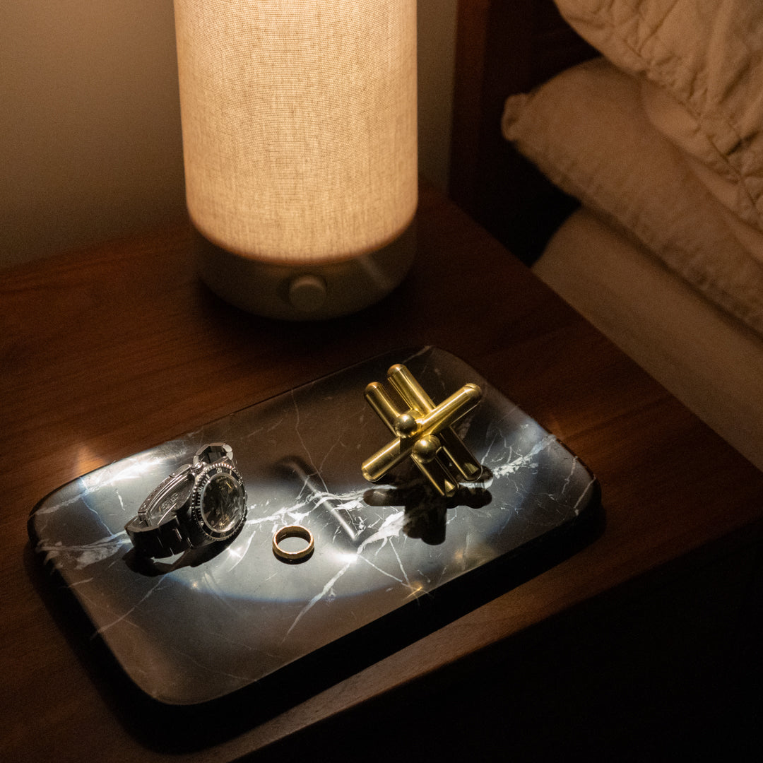 A dimly lit bedside table features a modern lamp, a dark marble tray with a wristwatch, a metallic ring, and a brass jack. The corner of a bed with white bedding is visible in the background.