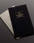 Two journals are stacked on a dark surface. The top journal, The Five Minute Journal, features elegant gold text on a black cover. A light beige notebook rests partially visible beneath it, creating a minimalist scene.