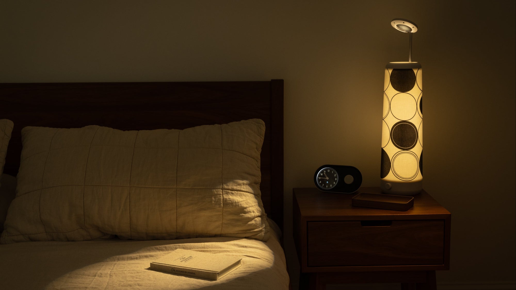 A softly lit bedroom scene with a bed, beige pillow, and a book on the covers. Beside the bed is a wooden nightstand holding a bedside lamp, an alarm clock, and a closed book. The lamp casts a bright reading spotlight.