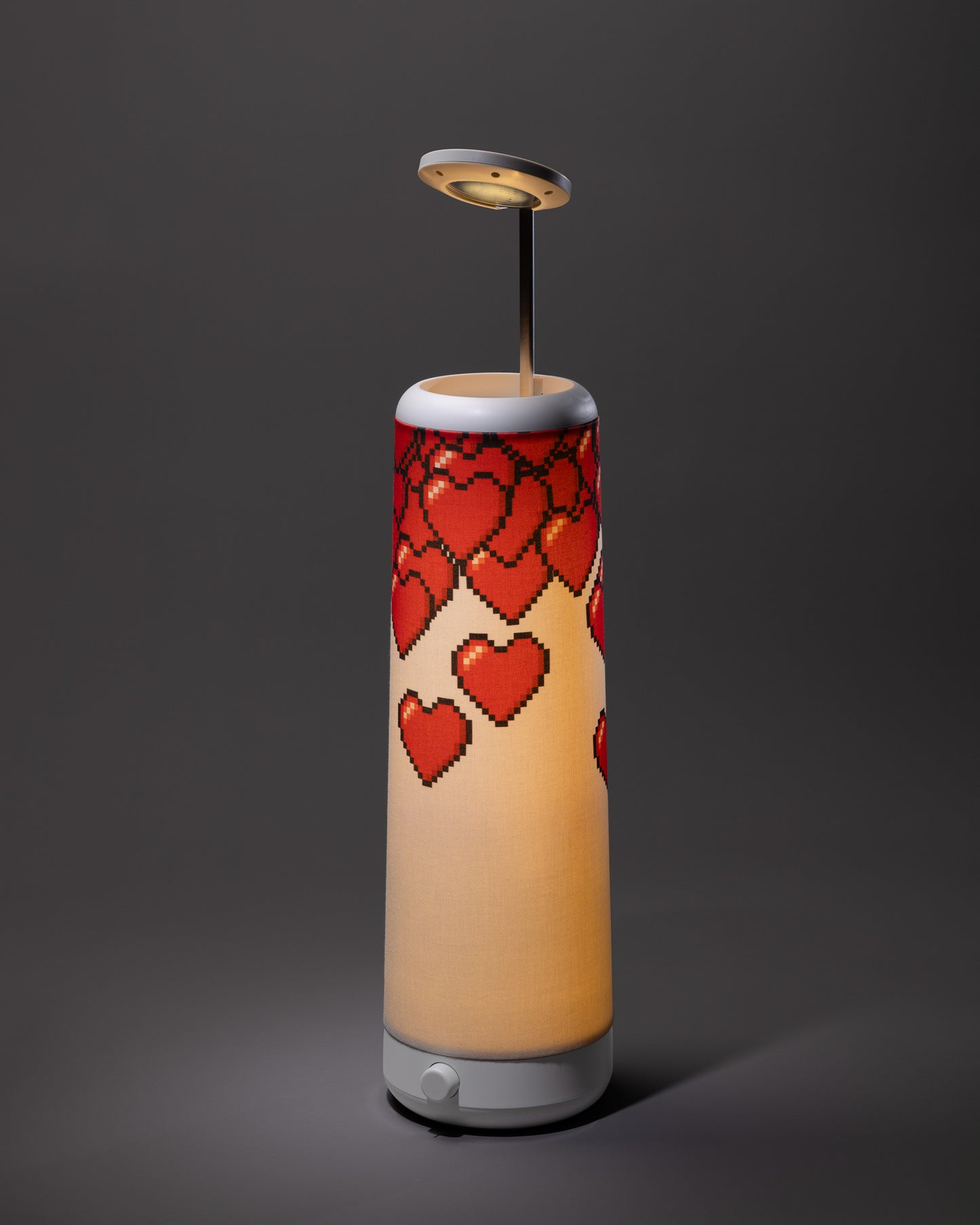 A cylindrical lamp with a white base and top, decorated with a pattern of red pixelated hearts. The lamp emits a warm light and has a modern, minimalist design against a dark gray background.