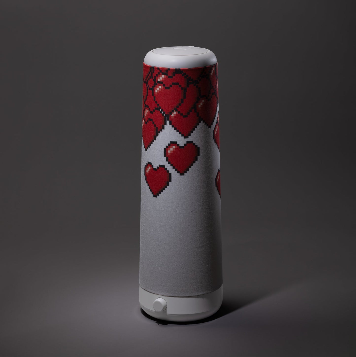A tall, cylindrical white table lamp decorated with pixelated red hearts on its upper half, set against a dark gray background.