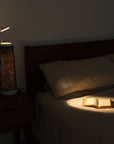 A cozy bedroom features a bed with beige plaid bedding and an open book under the constellation-themed lamp by Nightside on a wooden nightstand. Dim lighting, coupled with interchangeable fabric shades from the Starlight Cotton Blackout Shade collection, creates a warm ambiance.