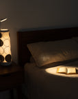 The bedroom features a bed with a beige quilt, softly lit by the Nightside lamps Concentric Circles Cotton Shade. Warm light highlights an open book on the bed, while blackout shades keep the room dim and cozy.