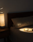 A cozy bedroom scene showcases a Nightside lamp with the Alphabet Cascade Cotton Shade, illuminating an open book on a neatly made bed. The warm glow highlights the pillow and partially lights up the wooden headboard and nightstand, with fabric shades offering versatile style.
