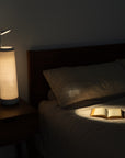 A cozy bedroom features a dimly lit Nightside lamp with a Cursive Lines Cotton Shade, illuminating an open book on the neatly made bed. The interchangeable fabric shades cast a soft glow, creating a warm and inviting atmosphere against the dark background.