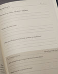 The Stoic Journal by Intelligent Change lies open, displaying morning and evening check-in prompts about intentions, control, mindset, interactions with others, and gratitude. The page is blank and ready to be filled in.