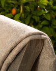 A close-up of the beige Midnight Sun Throw Blanket by Blacksaw, made from eco-friendly materials with visible stitching, is positioned in front of leafy green foliage and small orange fruits, with sunlight highlighting the texture.