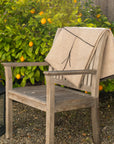 A wooden chair with the hypoallergenic Midnight Sun Throw Blanket by Blacksaw draped over it stands on gravel. A black lantern with a white candle sits nearby. Behind the chair, orange fruit adorns a leafy bush, and an eco-friendly wooden fence completes the scene.