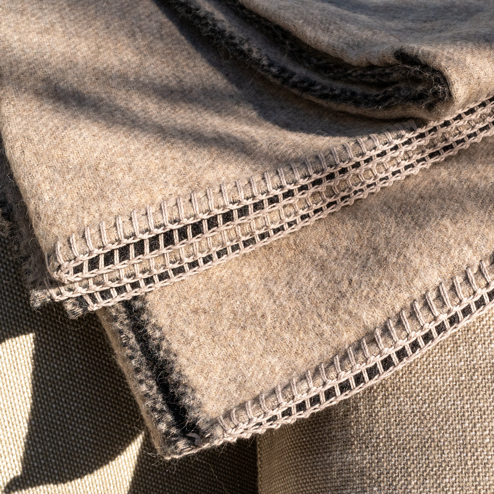 Close-up of folded Midnight Sun Throw Blankets by Blacksaw, made from eco-friendly baby alpaca. They feature intricate black and white stitching along the edges. The texture is soft and cozy, with sunlight casting gentle shadows over the hypoallergenic surface.