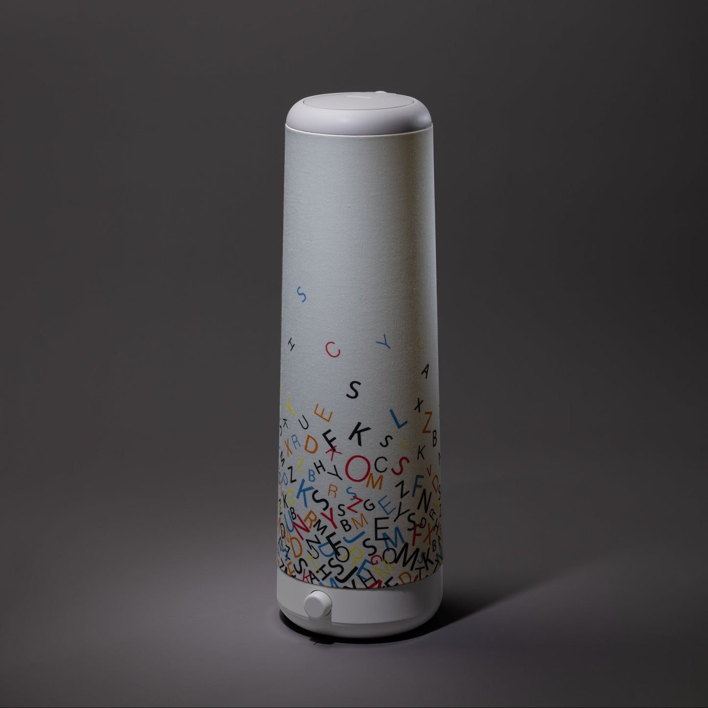 The Nightside Alphabet Cascade Cotton Shade lamp features a tall, cylindrical design with white accents and colorful alphabet letters, plus interchangeable fabric shades, displayed against a dark background.