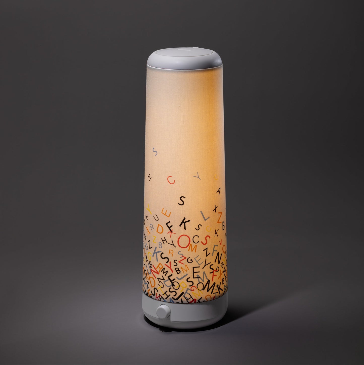 The Nightside Alphabet Cascade Cotton Shade is a modern cylindrical lamp with interchangeable fabric shades and a soft warm glow, featuring colorful scattered alphabet letters near its base against a dark grey background.