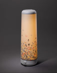 The Nightside Alphabet Cascade Cotton Shade is a modern cylindrical lamp with interchangeable fabric shades and a soft warm glow, featuring colorful scattered alphabet letters near its base against a dark grey background.