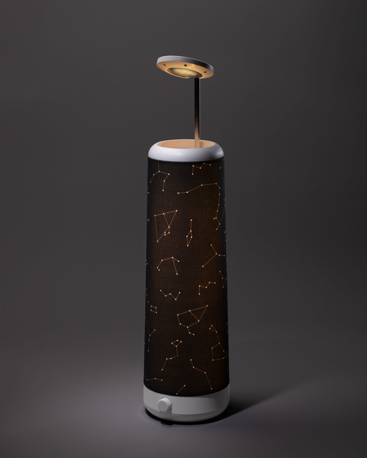 The Nightside Starlight Cotton Blackout Shade features illuminated star constellations on a cylindrical shade and stands on a white base, with a circular light source above, set against a dark background.