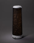 A cylindrical Nightside lamp with a Starlight Cotton Blackout Shade, featuring golden constellation patterns, stands on a plain gray surface against a gray background.