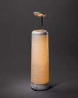 The Nightside Cursive Lines Cotton Shade lamp features wavy patterns and a soft yellow glow. It stands on a dark surface, topped by a small round fixture on a thin metal rod, with interchangeable fabric shades for versatile style.