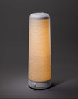 The Nightside Cursive Lines Cotton Shade lamp features a modern cylindrical design, a white base and top, interchangeable fabric shades with subtle wavy lines, and emits a soft, warm light against a dark background.