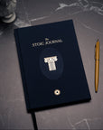 A dark navy blue Stoic Journal with gold lettering and a column emblem, shown closed on a dark marble surface next to a gold pen.
