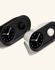 The Nanu Arc Alarm Clock comes in black and white, each with round black faces, white hands, green accents, a side knob, and minimalist wedge-shaped design on a light background evoking precise analog charm.