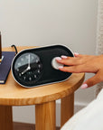 A persons hand gently reaches to turn off the Nanu Arc Alarm Clock on a wooden bedside table. The table also features a closed book, a gold decorative piece, and a lit candle in a brown glass holder.