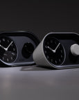Two Nanu Arc Alarm Clocks—one black, one white—sit side by side on a gray surface, both displaying 10:10. Each features a round knob, minimalist design, and convenient USB-C wall power for easy charging.