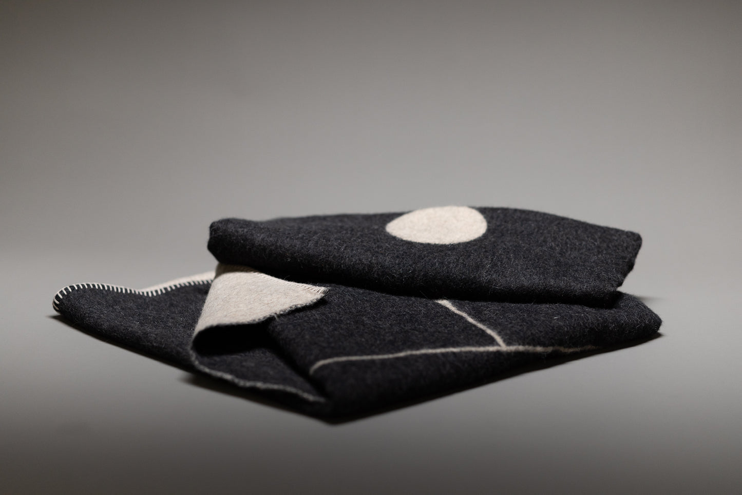 The Blacksaw Midnight Sun Throw Blanket is a folded dark wool Baby Alpaca blanket featuring a white circle and light geometric lines, shown against a gray background with a visible stitched seam on its hypoallergenic edge.