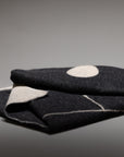The Blacksaw Midnight Sun Throw Blanket is a folded dark wool Baby Alpaca blanket featuring a white circle and light geometric lines, shown against a gray background with a visible stitched seam on its hypoallergenic edge.