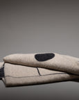 The Blacksaw Midnight Sun Throw Blanket is a folded beige wool piece with black stitched lines and a large black dot, made from eco-friendly sustainable fibers, shown on a plain gray background.