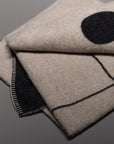 A close-up of the folded Midnight Sun Throw Blanket by Blacksaw, crafted from eco-friendly wool with black stitched edges and a minimal geometric pattern, shown on a smooth gray surface.