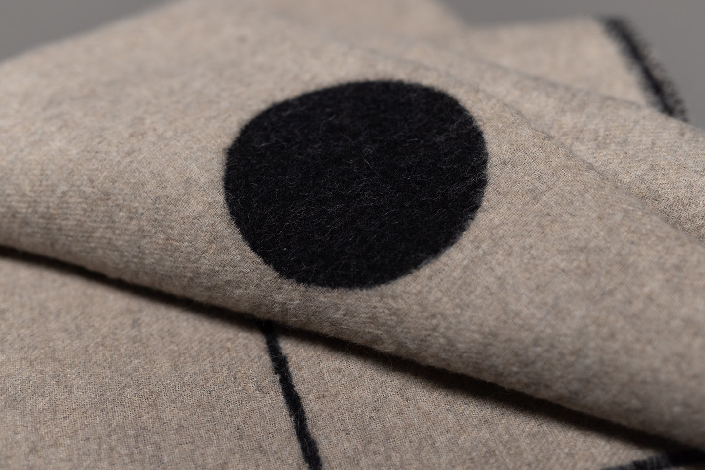 A close-up of the Midnight Sun Throw Blanket by Blacksaw, made from eco-friendly fibers in beige, featuring a large black circle and thin black line pattern. The folded fabric partially obscures the distinctive design.