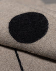 A close-up of the Midnight Sun Throw Blanket by Blacksaw, made from eco-friendly fibers in beige, featuring a large black circle and thin black line pattern. The folded fabric partially obscures the distinctive design.