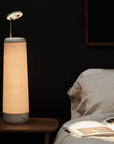 A tall, cylindrical Nightside NS01 Personal Task Lamp with a soothing Warm-White LED and a light brown shade sits on a wooden bedside table. Its gentle glow contrasts with the dark background wall, illuminating an open book on the bed beside a beige pillow.