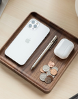 The Ugmonk Valet Tray - Medium, crafted from precision-milled walnut, sits on a light desk holding a silver iPhone, metallic pen, white wireless earbuds in a case, and assorted coins—showcasing its unique grain and elegant design.