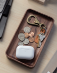 On the desk, the Ugmonk Valet Tray - Small, crafted from solid walnut, organizes coins, a brass bottle opener, two keys on a leather keychain, and a white wireless earbuds case. A gray sleeve and other items are nearby.