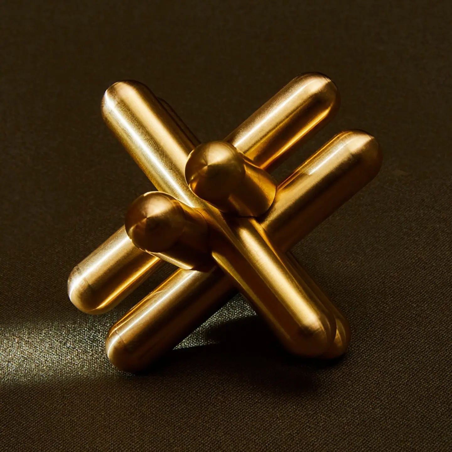 The Brass Jack Puzzle by Craighill is a gold metallic sculpture crafted from solid brass, resembling interlocking rods. Measuring 3x3x3 with a 1/2 diameter, it sits on a dark textured surface, reflecting light off its smooth, shiny finish and highlighting its three-dimensional shape.