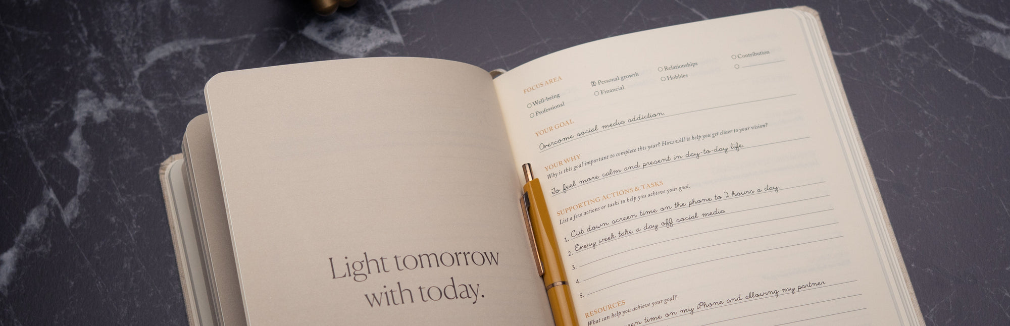 An open journal lies on a dark marble surface, featuring a gold pen. One page reads Light tomorrow with today, while the other has prompts for priorities, gratitude, and schedule planning.