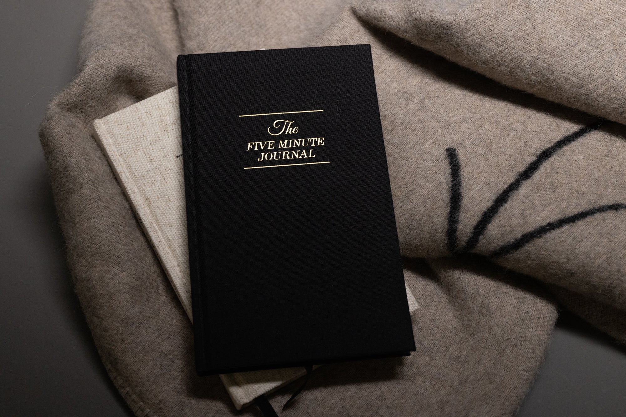 A black hardcover book titled The Five Minute Journal rests on top of a light-colored book and a soft beige blanket.