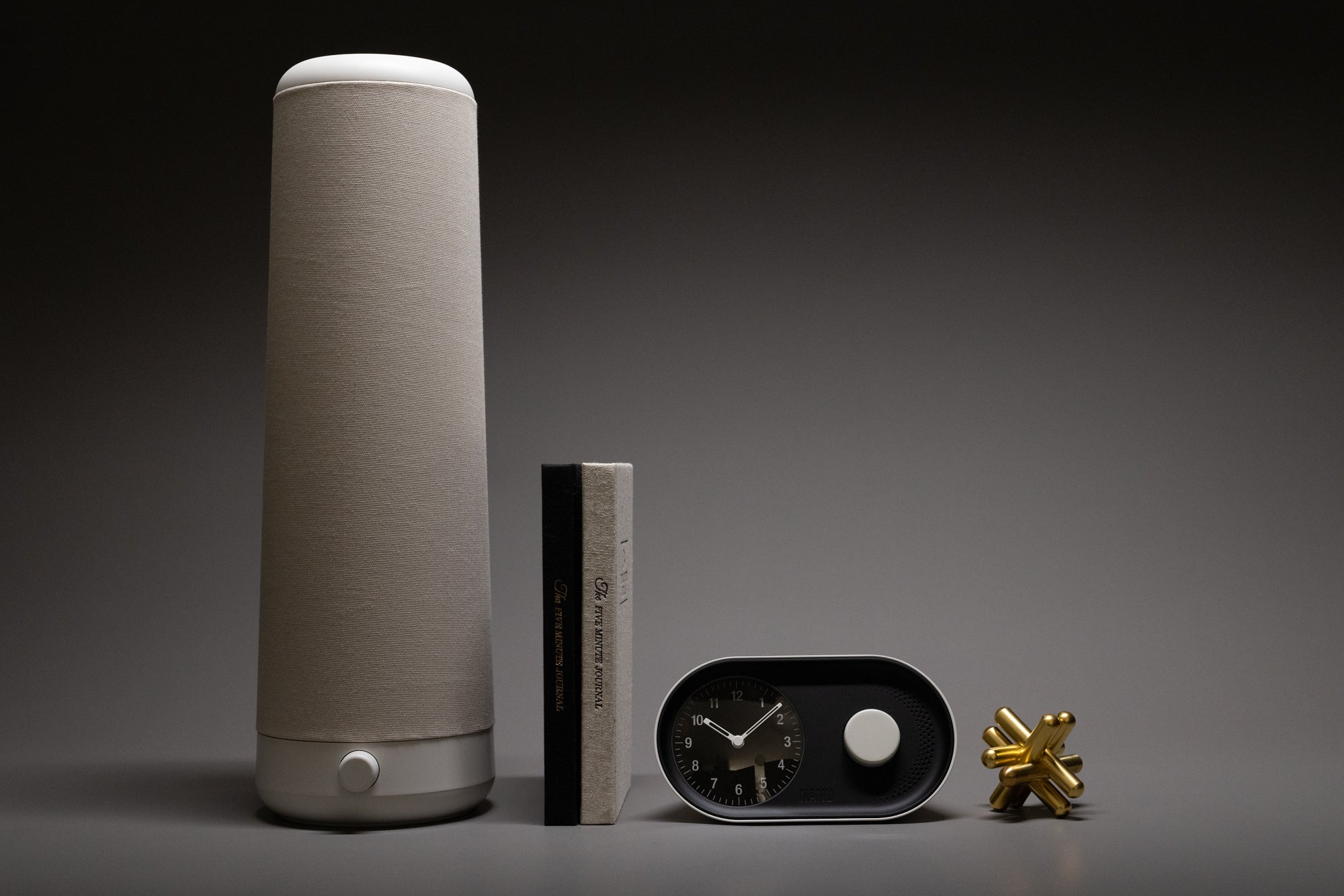 A tall, fabric-covered smart speaker, two stacked books, a small modern black clock, and a gold jacks game piece arranged against a gray background.