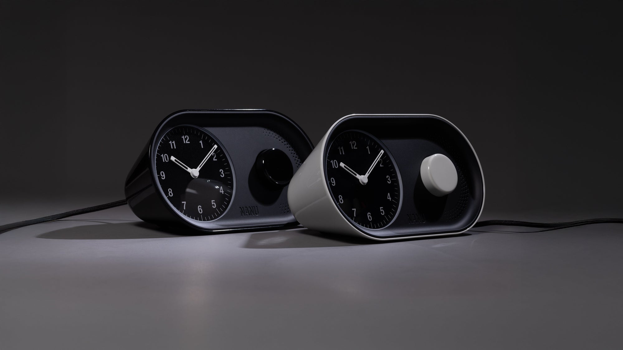 Two modern, minimalist analog clocks, one black and one white, are placed side by side on a grey surface with a dark background. Both clocks feature rounded, rectangular shapes and simple dials.