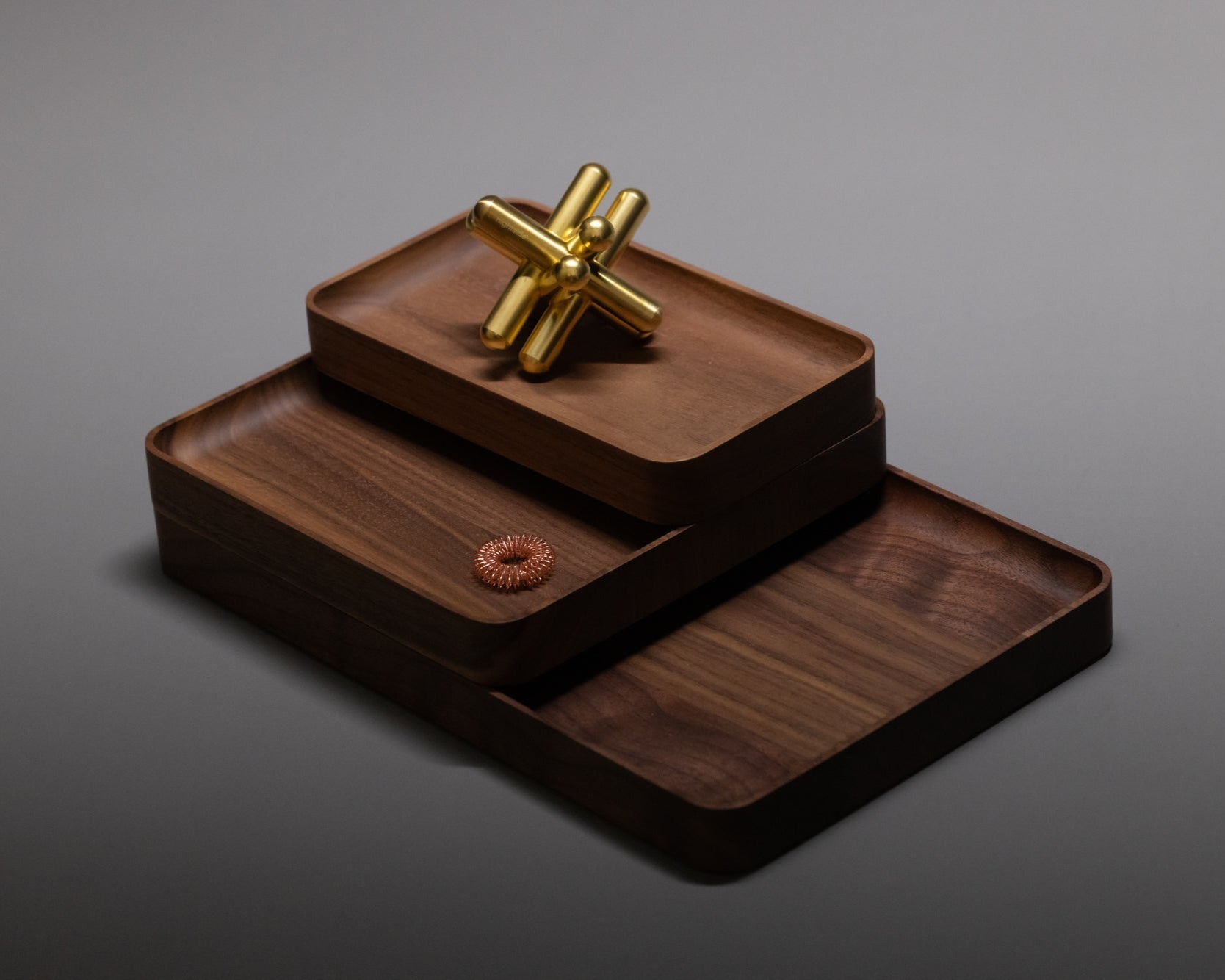 Three stacked rectangular wooden trays hold a gold metal jack and a copper hair tie, all placed on a smooth grey surface with soft lighting.
